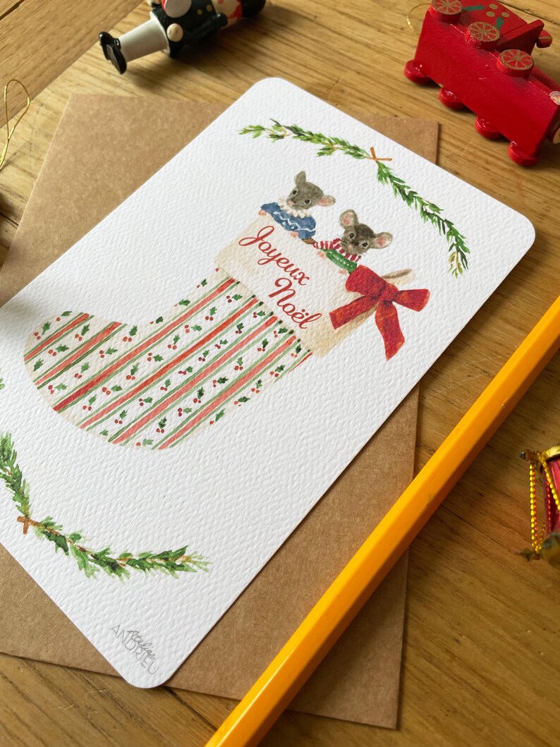 Puede incluir: Tarjeta de Navidad con una ilustraci&oacute;n en acuarela de un calcet&iacute;n y dos ratones. El calcet&iacute;n tiene rayas y bayas rojas y verdes, con las palabras "Joyeux No&euml;l". La tarjeta est&aacute; sobre un sobre marr&oacute;n.