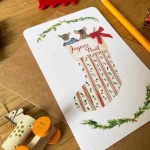 Puede incluir: Una tarjeta de Navidad con un dise&ntilde;o de calcet&iacute;n y dos ratones. La tarjeta tiene el texto "Joyeux No&euml;l" y tiene rayas y ramitas rojas y verdes. Tambi&eacute;n se ve un tren de madera rojo y un l&aacute;piz.