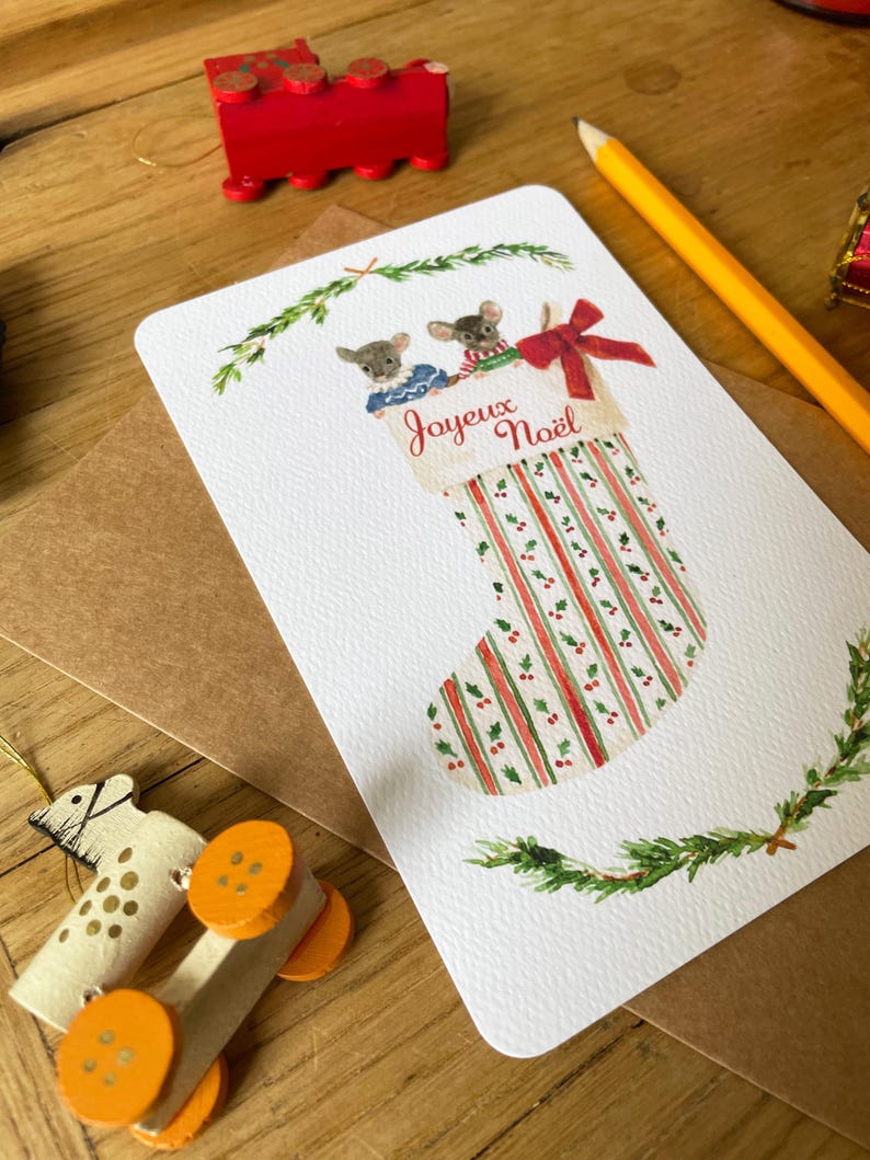 Puede incluir: Una tarjeta de Navidad con un dise&ntilde;o de calcet&iacute;n y dos ratones. La tarjeta tiene el texto "Joyeux No&euml;l" y tiene rayas y ramitas rojas y verdes. Tambi&eacute;n se ve un tren de madera rojo y un l&aacute;piz.