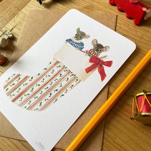 Puede incluir: Tarjeta de Navidad con una ilustraci&oacute;n en acuarela de dos ratones asom&aacute;ndose por una media. La media es blanca con rayas rojas y verdes y un lazo rojo. Otras decoraciones navide&ntilde;as se ven en el fondo.