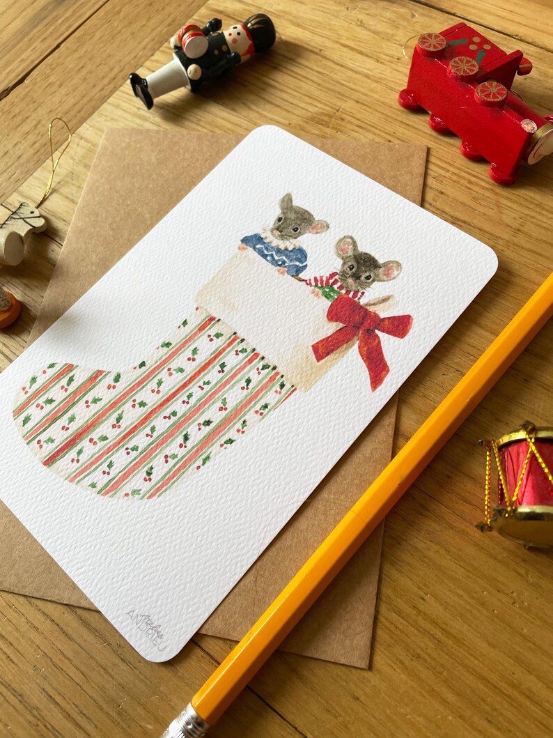 Puede incluir: Tarjeta de Navidad con una ilustraci&oacute;n en acuarela de dos ratones asom&aacute;ndose por una media. La media es blanca con rayas rojas y verdes y un lazo rojo. Otras decoraciones navide&ntilde;as se ven en el fondo.