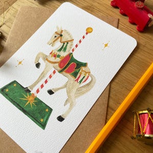 以下が含まれることがあります： 白いメリーゴーランドの馬のイラストが描かれたグリーティングカード。馬は緑と赤の鞍を持ち、白い背景に配置されています。赤と白のストライプのポールが馬から伸びています。カードは木製の表面にあります。