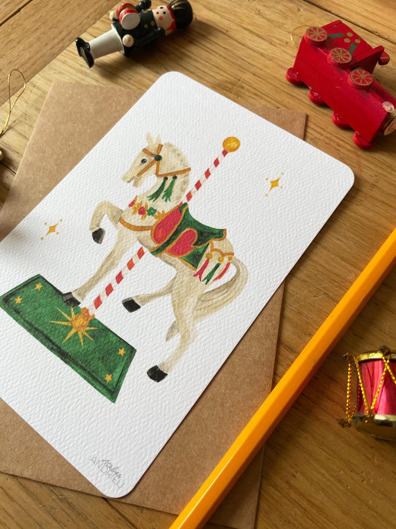 以下が含まれることがあります： 白いメリーゴーランドの馬のイラストが描かれたグリーティングカード。馬は緑と赤の鞍を持ち、白い背景に配置されています。赤と白のストライプのポールが馬から伸びています。カードは木製の表面にあります。