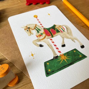 以下が含まれることがあります： 長方形のカードに描かれた白いメリーゴーランドの馬のイラスト。馬は緑色の鞍と赤いアクセントがあります。カードは木製の表面にあり、赤いおもちゃの電車と黄色の鉛筆があります。