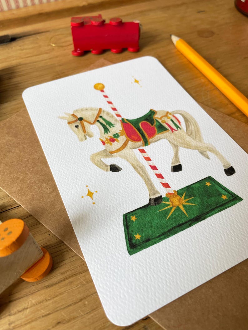 以下が含まれることがあります： 長方形のカードに描かれた白いメリーゴーランドの馬のイラスト。馬は緑色の鞍と赤いアクセントがあります。カードは木製の表面にあり、赤いおもちゃの電車と黄色の鉛筆があります。