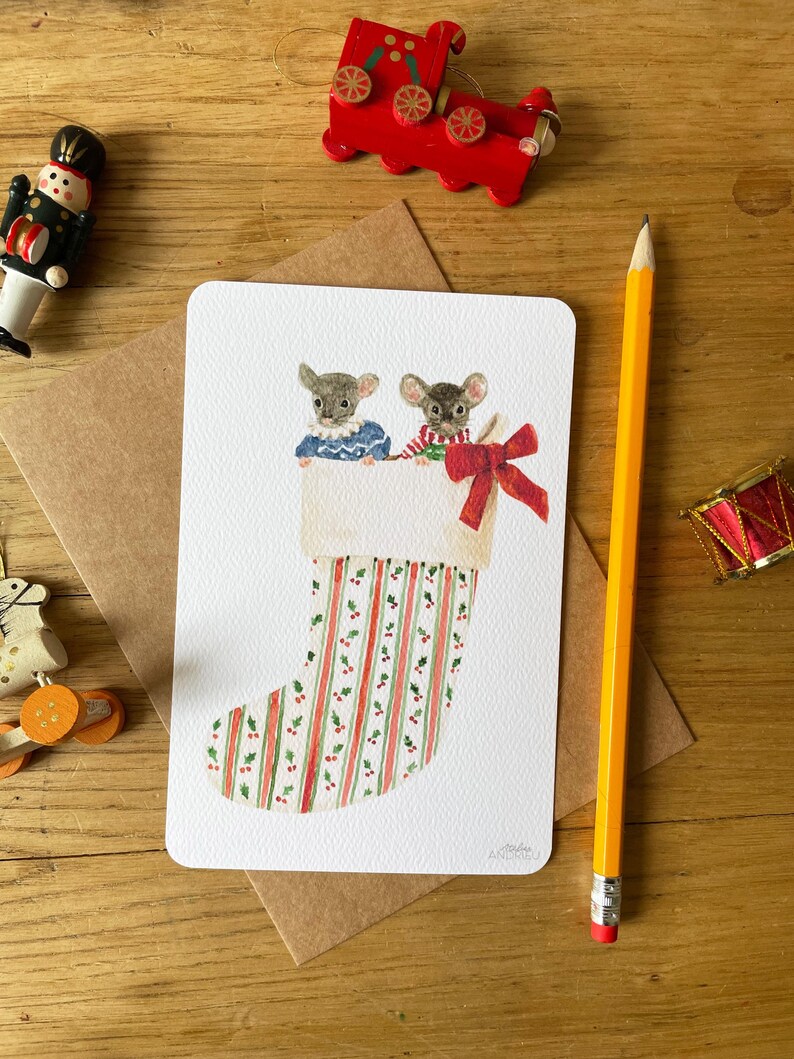 Puede incluir: Una tarjeta de Navidad con dos ratones mirando por encima de un calcet&iacute;n festivo. El calcet&iacute;n tiene rayas rojas y verdes y acebo. Un lazo rojo est&aacute; atado en la parte superior. Un tren de madera rojo y otras decoraciones est&aacute;n en el fondo.