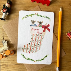 Puede incluir: Una tarjeta de Navidad con una ilustraci&oacute;n en acuarela de dos ratones asom&aacute;ndose por una bota navide&ntilde;a. La bota est&aacute; decorada con rayas rojas y verdes y lleva la inscripci&oacute;n "Joyeux No&euml;l". Un lazo rojo adorna la bota. Decoraciones navide&ntilde;as rodean la tarjeta.