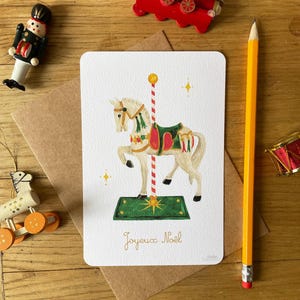 以下が含まれることがあります： メリーゴーランドの馬のイラストが描かれたクリスマスカード。カードの下には「Joyeux No&euml;l」の文字。カードの周りには、おもちゃの電車、くるみ割り人形、鉛筆など、クリスマスの飾り付けが施されている。