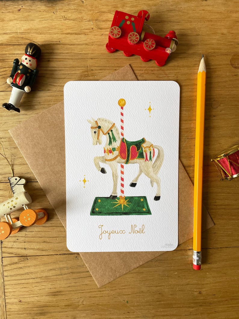 以下が含まれることがあります： メリーゴーランドの馬のイラストが描かれたクリスマスカード。カードの下には「Joyeux No&euml;l」の文字。カードの周りには、おもちゃの電車、くるみ割り人形、鉛筆など、クリスマスの飾り付けが施されている。