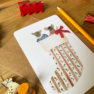Puede incluir: Una tarjeta navide&ntilde;a con dos ratones asom&aacute;ndose de un calcet&iacute;n navide&ntilde;o. El calcet&iacute;n tiene rayas rojas y verdes con detalles de acebo. Un tren de madera rojo y otras decoraciones navide&ntilde;as est&aacute;n en el fondo.