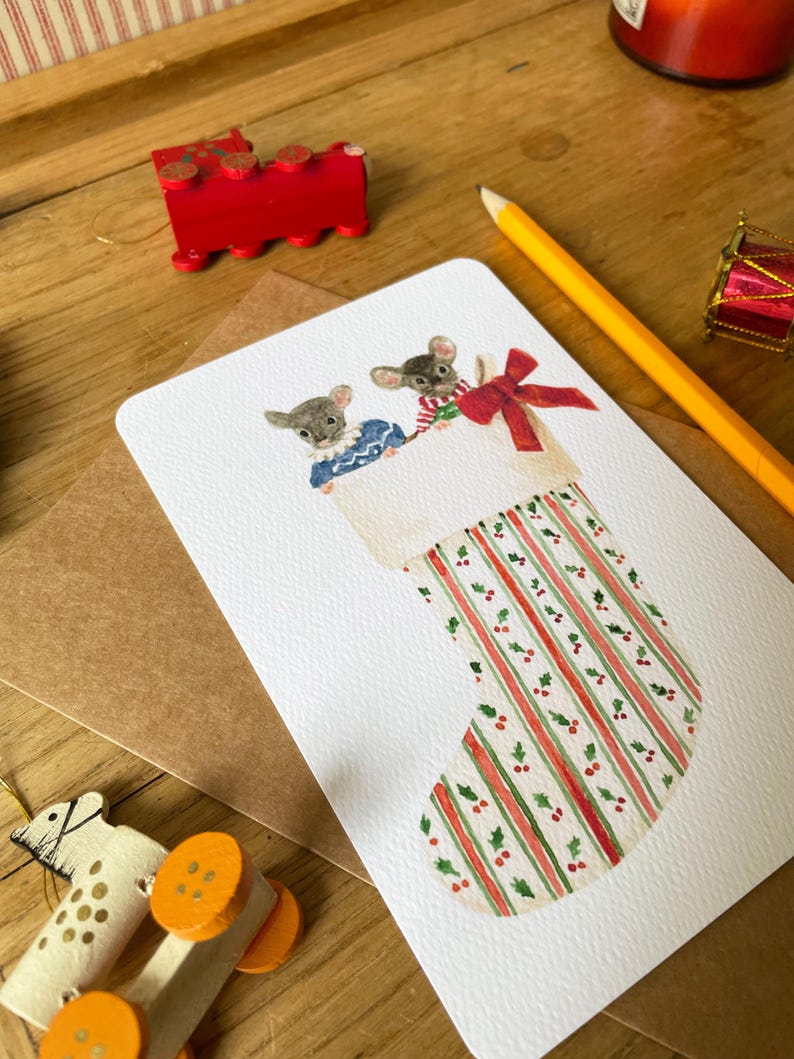 Puede incluir: Una tarjeta navide&ntilde;a con dos ratones asom&aacute;ndose de un calcet&iacute;n navide&ntilde;o. El calcet&iacute;n tiene rayas rojas y verdes con detalles de acebo. Un tren de madera rojo y otras decoraciones navide&ntilde;as est&aacute;n en el fondo.