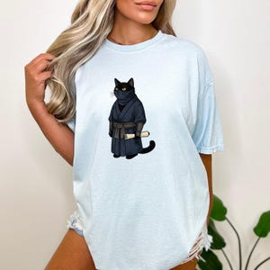 May include: A light blue short-sleeve t-shirt featuring a graphic illustration of a black cat dressed as a ninja. The cat wears a dark blue robe with a matching belt and holds a small, rolled-up parchment scroll in its front paws.