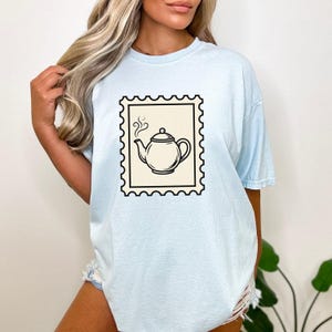 Könnte beinhalten: Ein hellblaues Kurzarm-T-Shirt mit einer zentralen Grafik in Form einer cremefarbenen Briefmarke. Die Briefmarke zeigt eine schwarze Strichzeichnung einer Teekanne mit aufsteigendem Dampf, umrahmt von einem gezackten schwarzen Rand.