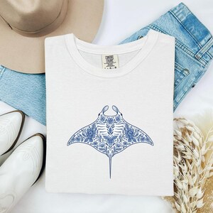 Könnte beinhalten: Weißes T-Shirt mit blauem, floralem Mantarochen-Design. Die Illustration zeigt Blumen und Muster. Das gefaltete Shirt wird mit Jeans, Stiefeln und einem Hut präsentiert.