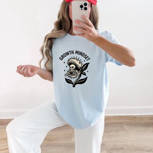 May include: A light blue t-shirt featuring a black graphic print of a Venus flytrap plant with a skeleton inside making a heart shape with its hands. The text "GROWTH MINDSET" is printed in a black arched font above the plant illustration.