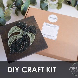May include: A DIY craft kit featuring a string art project of a small monstera leaf. The leaf design is made with dark green and light green string on a dark brown wooden square. The kit includes a brown cardboard box with the text "STRING ART KIT".