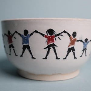 May include: White ceramic bowl with a painted design of children holding hands in a circle. The children are depicted in black silhouettes, wearing red, blue, and orange shirts. The bowl has a slightly flared shape.