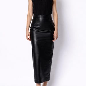 Puede incluir: Un top negro con cuello halter y una falda lápiz de cuero negro. La falda llega a la rodilla. La modelo lleva sandalias negras con tiras. El conjunto está sobre un fondo blanco.