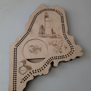 May include: Wooden cribbage board in the shape of the state of Maine. The board features engraved images of a lighthouse, moose, lobster, and a loon. The words "Start" and "Finish" are also engraved on the board.