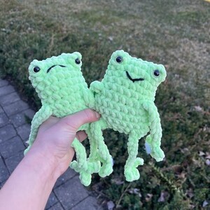 May include: Two green crocheted frog plushies with black eyes and smiling mouths. The frogs are sitting on a hand.