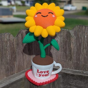May include: A smiling yellow sunflower with a brown center and green leaves is in a white teacup with a heart-shaped handle. The teacup is on a pink heart-shaped coaster with red trim. The text "Love you!" is on the front of the teacup.