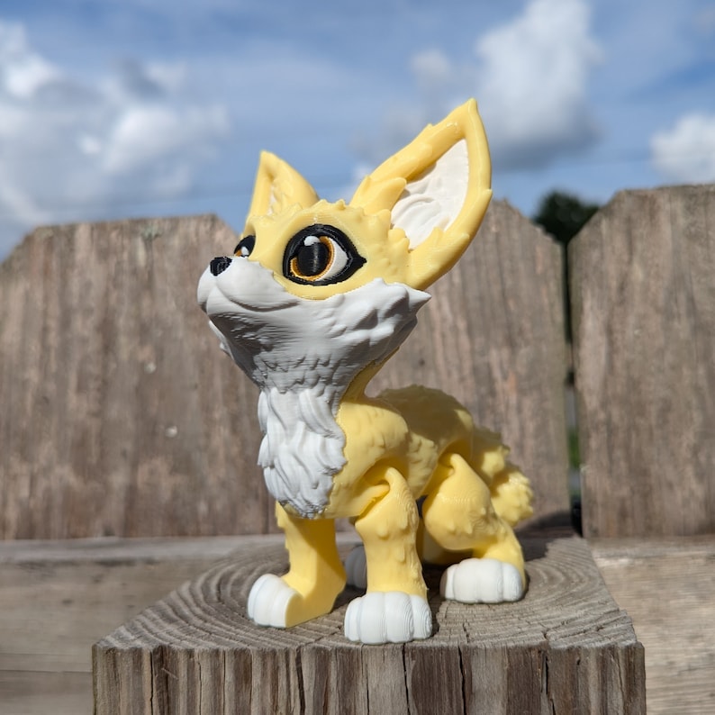 A cute 3D printed fennec fox stands on a fence post. It is yellow and white with black and gold eyes. It has huge ears, big eyes, and a clever look on its face.