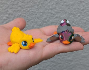May include: Two small, colorful duck figurines. One is bright yellow with orange accents and blue eyes. The other is a mix of gray, purple, brown, and orange, with large eyes. Both are resting on a hand.