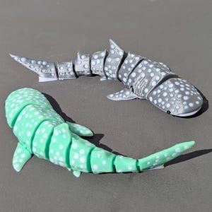 May include: Two articulated whale shark toys. One is light gray with white spots, the other is mint green with white spots. The toys are segmented, allowing for movement. The toys are designed to resemble whale sharks.