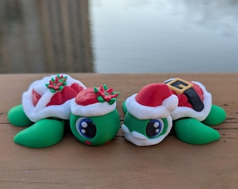 May include: Two green turtle figurines with festive Santa Claus hats and outfits. One turtle has a red and white shell with holly accents, while the other wears a Santa suit. The turtles are on a wooden surface.