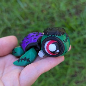 May include: A 3D-printed turtle figurine with a Frankenstein-inspired design. The turtle has a green head and limbs, a purple shell, and a black top. The eye is large with red, white, and black details. The figurine is held in a hand.
