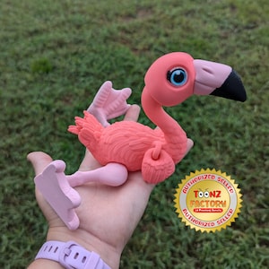 May include: Pink flamingo toy with blue eyes and a black beak. The toy is articulated and has a pink and white color scheme. The toy is held in a hand with a purple wristband. The image also includes a gold sticker with the text "AUTHORIZED SELLER TOONZ FACTORY 12 Printing Service AUTHORIZED SELLER".