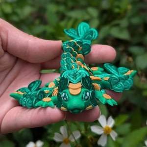 May include: A green and orange 3D printed dragon figurine with a leafy design. The dragon has large eyes and a small mouth.