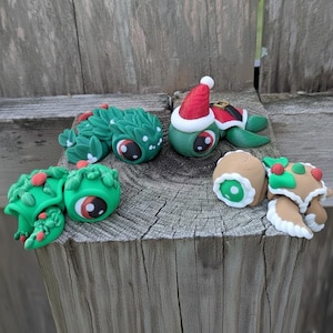 May include: A collection of whimsical, handmade clay figurines. The figurines include a green lizard-like creature, a turtle wearing a Santa hat, and several round, eye-shaped figures with festive decorations. The color palette is primarily green, red, and brown.