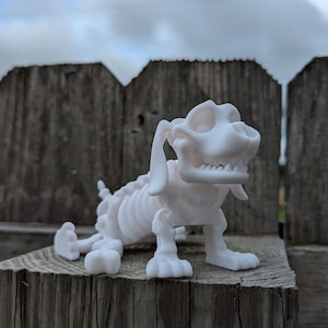 May include: A white plastic skeleton dog figurine with a playful expression. The dog is posed sitting up with its front paws extended and its back legs tucked underneath. The figurine is made of a smooth, white plastic and has a detailed skeletal structure.