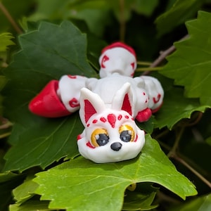 May include: A white and red fox figurine with large eyes and a decorative pattern. The fox is surrounded by green leaves. The figurine has a unique design and is likely a handmade craft item.