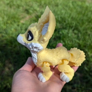 A cute 3D printed fennec fox is held in a hand. It is yellow and white with black and gold eyes. It has huge ears, big eyes, and a clever look on its face.