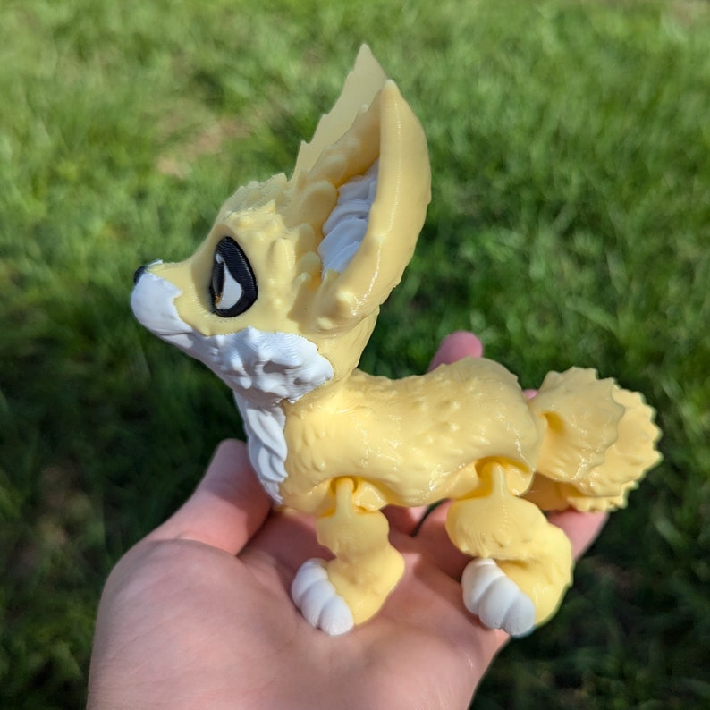 A cute 3D printed fennec fox is held in a hand. It is yellow and white with black and gold eyes. It has huge ears, big eyes, and a clever look on its face.