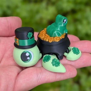 May include: A whimsical St. Patrick's Day-themed figurine set. Features a light green eye-shaped character wearing a black top hat with a green band and shamrock, and a frog atop a pot of gold. The set includes green shamrock-adorned feet.