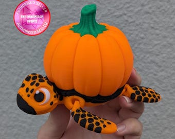 May include: A 3D printed orange pumpkin with a green stem sits on top of a black and orange turtle. The turtle has a large, round eye and a small mouth.