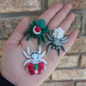 May include: Three small, hand-painted, Christmas-themed spider ornaments. One is green with a red Santa hat, one is white with a red bow, and one is gray with a white fluffy top.