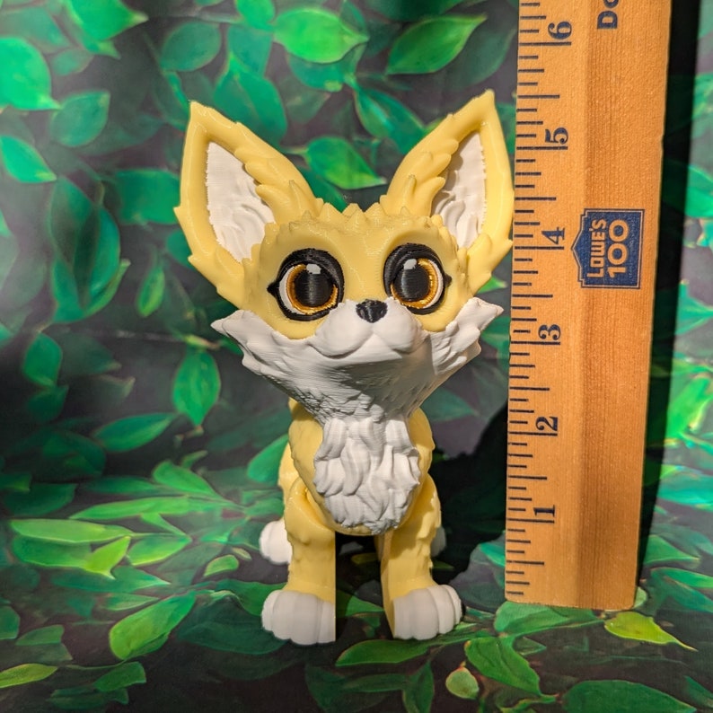A cute 3D printed fennec fox stands against a forest backdrop. It is yellow and white with black and gold eyes. It has huge ears, big eyes, and a clever look on its face. It is approximately 5.5 inches tall.