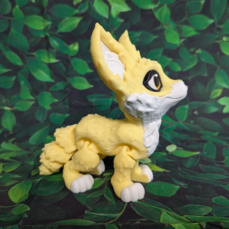 A cute 3D printed fennec fox stands against a forest backdrop. It is yellow and white with black and gold eyes. It has huge ears, big eyes, and a clever look on its face.