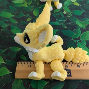 A cute 3D printed fennec fox stands against a forest backdrop. It is yellow and white with black and gold eyes. It has huge ears, big eyes, and a clever look on its face. It is approximately 6 inches long.