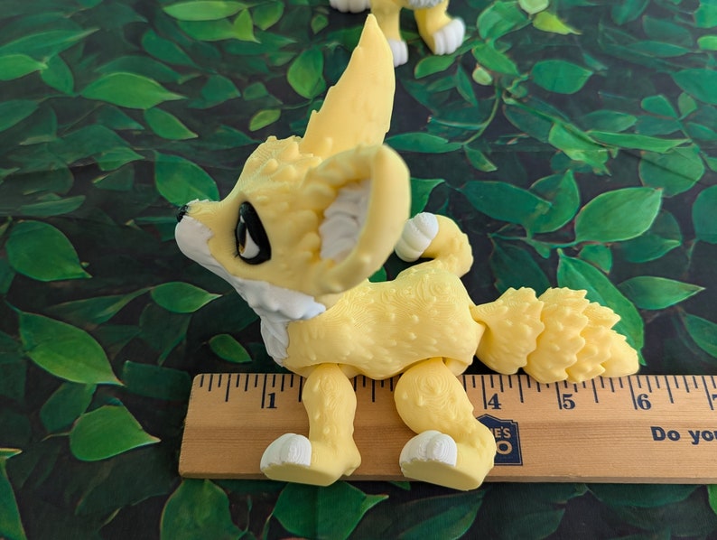 A cute 3D printed fennec fox stands against a forest backdrop. It is yellow and white with black and gold eyes. It has huge ears, big eyes, and a clever look on its face. It is approximately 6 inches long.