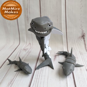 May include: Three gray articulated shark figurines on a woodgrain background. The largest shark has a white underside and a toothy grin. The text "MatMire Makes" is visible on a brown circular sticker in the upper left corner.