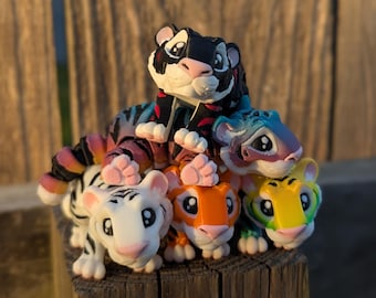 May include: A stack of five small tiger figurines. The top tiger is black with red and white stripes. The next tiger is blue with white stripes. The third tiger is white with black stripes. The fourth tiger is orange with black stripes. The bottom tiger is yellow with black stripes.
