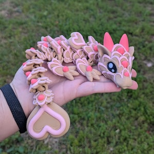 May include: A pink and brown dragon figurine with a heart-shaped tail. The dragon has pink accents and a detailed design.