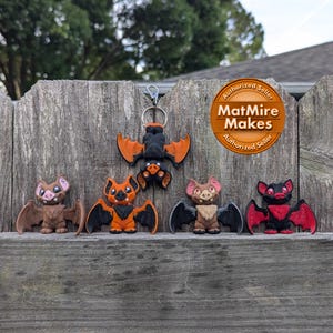 May include: Five handmade bat figurines, each with unique color combinations. The bats are brown, orange, red, and black. The bats are arranged in a row on a wooden surface. The bats are all in a flying position with their wings spread out. The bats are all about 2 inches tall. The bats are all made of polymer clay. The bats are all hand-painted. The bats are all unique and one-of-a-kind. The bats are all perfect for Halloween decorations or gifts. The bats are all made by MatMire Makes.