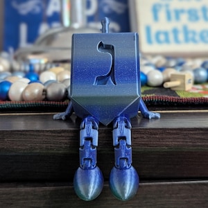 May include: A blue, glittery, articulated dreidel figure with a Hebrew letter cutout on its front face. The figure features a square body, a small top stem, and two jointed, dangling legs with rounded feet, all in a shimmering, gradient blue finish.