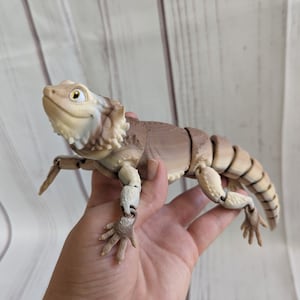 May include: A beige and brown articulated toy lizard with a spiky head and a friendly expression. The lizard is posed with its front legs raised and its back legs extended.
